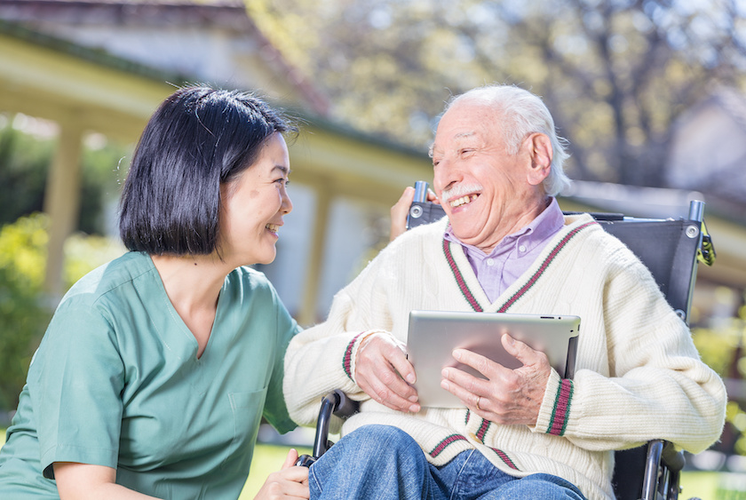 Happy elder patient in rehab clinic garden using tablet and smiling with nurse.